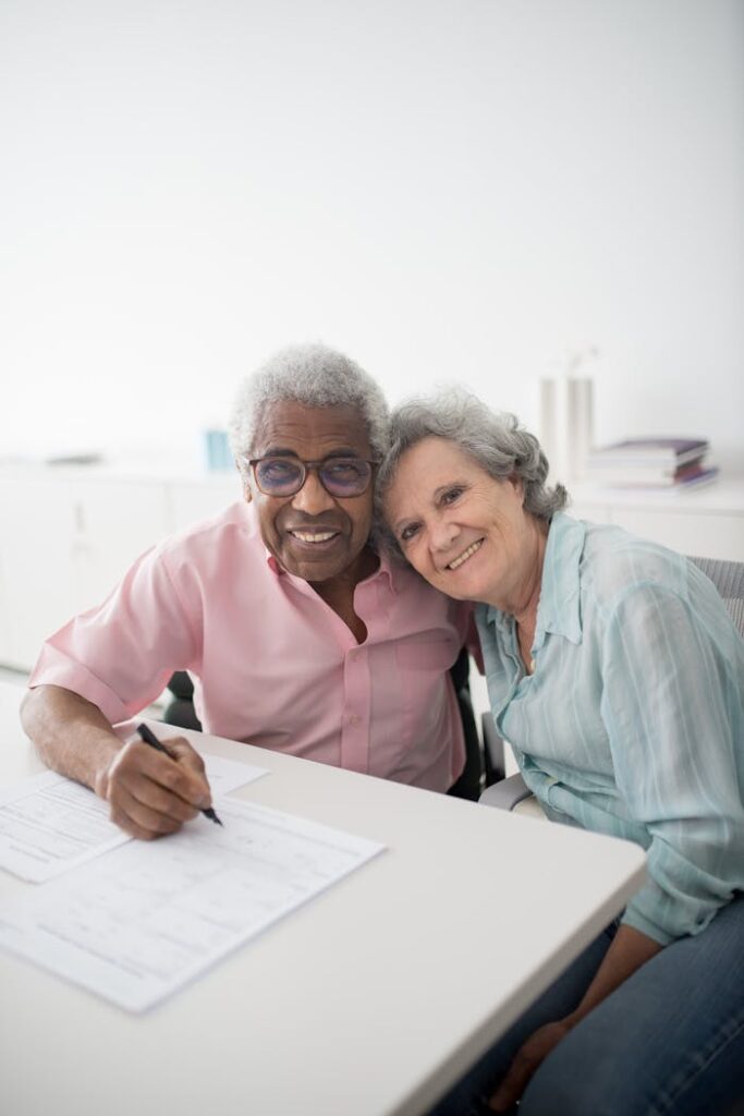 pexels-photo-8441866 Happy elderly couple sitting at a desk, smiling and signing documents. Perfect for insurance and lifestyle themes.